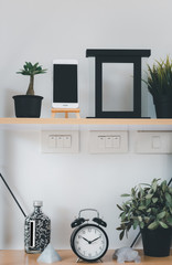 Wooden shelf with mobile phone,grass in pot, greenery in vase, alarm,photo frame and black board  over white wall  decoration in living room at home
