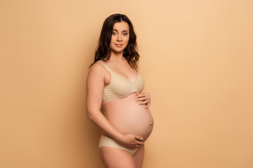  pregnant woman in lingerie looking at camera while touching tummy on beige