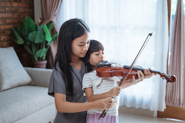 teenage girls help a child hold the violin bow properly while playing the violin indoors against the background of the window