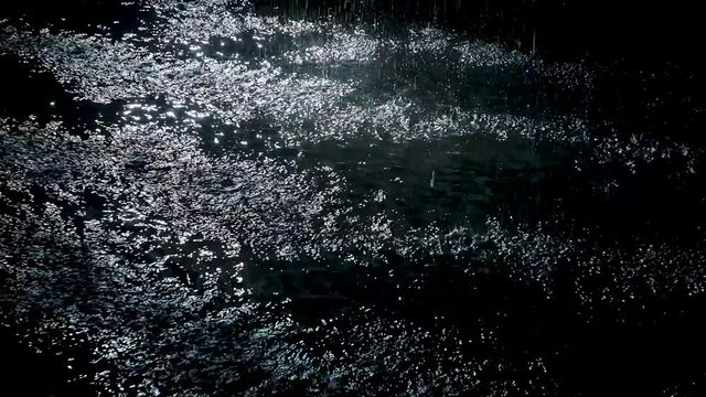 Rain on black. Spatters fall against on the water surface which glow. White neon lighting in dark studio. Slow motion. Close up.