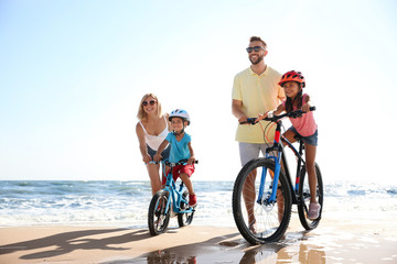 Happy parents teaching children to ride bicycles on sandy beach near sea