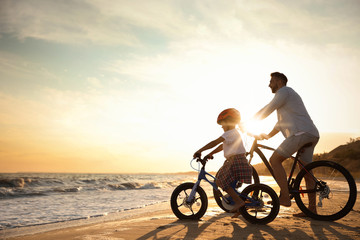 Fototapeta na wymiar Happy father with son riding bicycles on sandy beach near sea at sunset