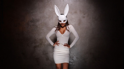 Portrait of beautiful flirting Asian woman in white dress and rabbit mask playfully posing for camera in the spotlight