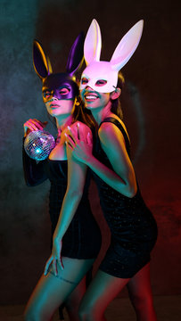 Portrait of two young flirting cute Asian women in black and white rabbit mask playfully posing for camera with small disco ball on the neon lights