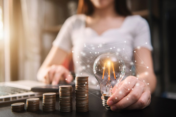 Business woman hand holding lightbulb with coins stack on desk. concept saving energy and money at office.