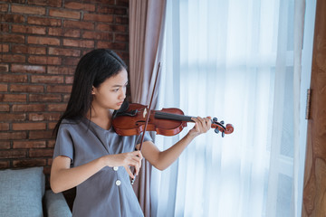 teenage girl learning to play a violin instrument in a room at home against a curtain background