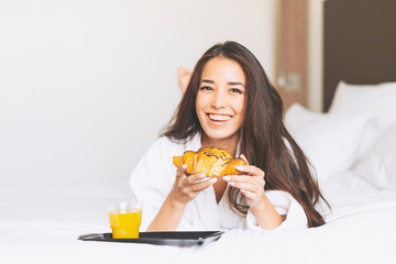 Beautiful happy Asian girl with long hair in white robe having breakfast Croissant and orange juice in the bed of hotel room
