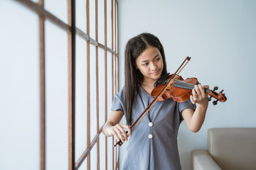 teenage girl learn to play a violin instrument beside the window in a room at home