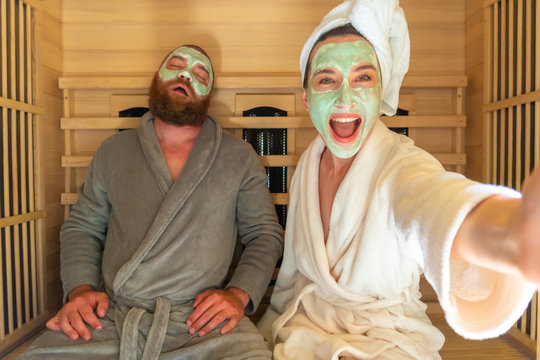 A Young Funny Couple Having Relax SPA Day In Sauna, A Woman Making Selfie With Beauty Mask While Her Boyfriend Is Asleep. Home Self Care, Body Care, Peace Of Mind And Infrared Sauna Concept.