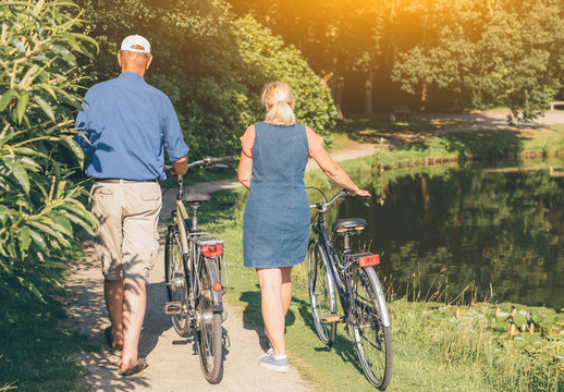 Back view of senior couple walking with bicycles in the park at sunset. Concept of active old people, healthy and sustainable lifestyle, ecological transport.