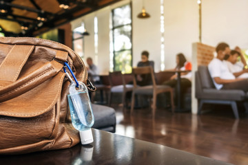 Fototapeta na wymiar Mini portable alcohol gel bottle to kill Corona Virus(Covid-19) hang on a brown leather shoulder bag on table in coffee shop.New normal lifestyle. Health care concept. Selective focus on alcohol gel