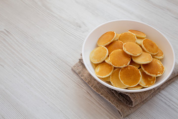 Homemade Mini Pancakes Cereal in a white bowl on a white wooden background, side view. Copy space.