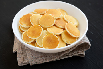 Homemade Mini Pancakes Cereal in a white bowl on a black surface, side view. Close-up.