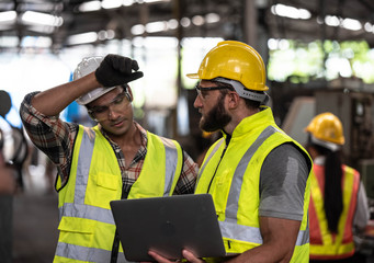 Two industrial Engineers and Technician workers with helmet or hardhat working and checking machinery on notebook or laptop in industry manufacturing Factory