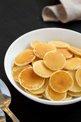 Homemade Mini Pancakes Cereal in a white bowl on a black background, side view.