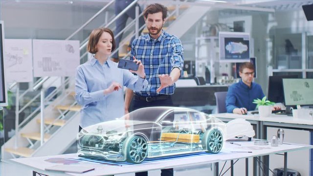 Industrial Design: Automotive Engineer and Designer Working on 3D Electric Car Design, Using Smartphone with Augmented Reality. Graphical Engine, Battery, Chassis, Body Collect into Full Vehicle