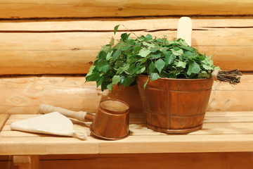 A fresh birch broom and copper vat and ladle are on a wooden bench in sauna.
