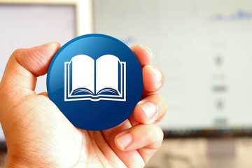 Book icon blue round button holding by hand infront of workspace background