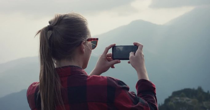 Young woman on a hike wear sunglasses take photo with her smartphone cell phone or mobile hiking in the autumn or fall clouds over mountains over shoulder slowmotion
