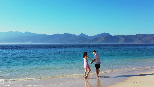 Wide shot of a lovely couple dancing at the beach during their intimate honeymoon near the waving sea with a mountain range on the horizon under the blue sky.