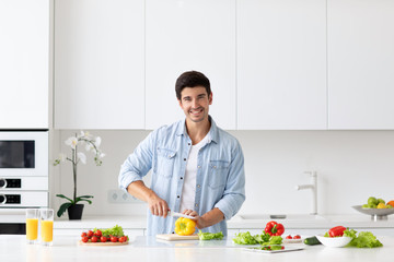 Young man cutting vegetables for salad at table in modern light kitchen.
