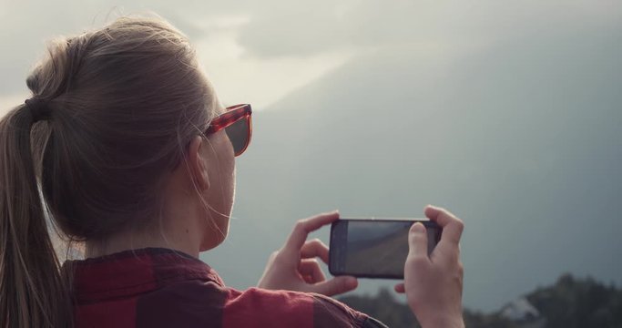 Young woman on a hike wear sunglasses take photo with her smartphone cell phone or mobile hiking in autumn or fall clouds over mountains over shoulder slowmotion close up