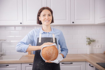 Woman baker in apron holds round bread in hands on background of white kitchen in Scandinavian style