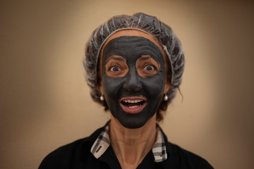 Brazilian woman with happy expression and gray clay mask. Concept: beauty, face care, facelift, wellness, skin care. Shot with brown background
