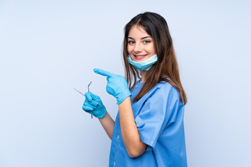 Woman dentist holding tools over isolated blue background pointing finger to the side