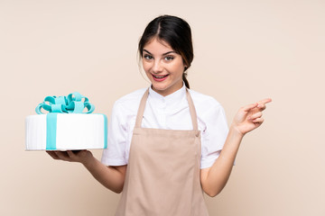 Pastry chef woman holding a big cake over isolated background surprised and pointing finger to the side