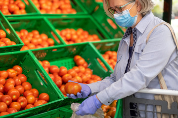 Senior woman wearing surgical mask and rubber gloves shopping and choosing fresh tomatoes in supermarket, healthy eating, active retired elderly people