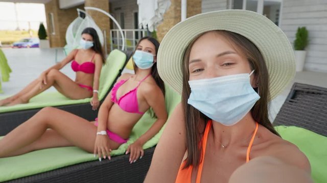 quarantine summer selfie, merry young girlfriends with protective medical masks observe safety rules while relaxing on sun loungers by pool with smartphone