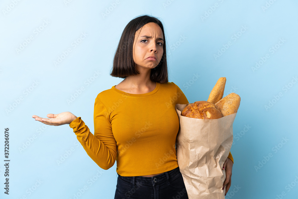 Wall mural Young latin woman buying some breads isolated on blue background making doubts gesture