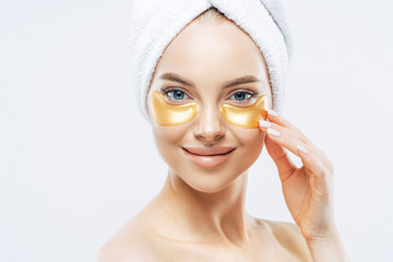 Close up shot of happy tender woman applies golden patches under eyes, removes wrinkles and dark circles, has manicure, wears towel on head, isolated over white background. Beauty treatment concept