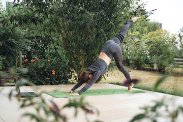 Active young woman in three legged downward dog pose on exercise mat with laptop in backyard