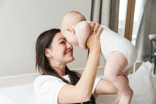 Photo of young excited mother smiling and playing with her baby on bed