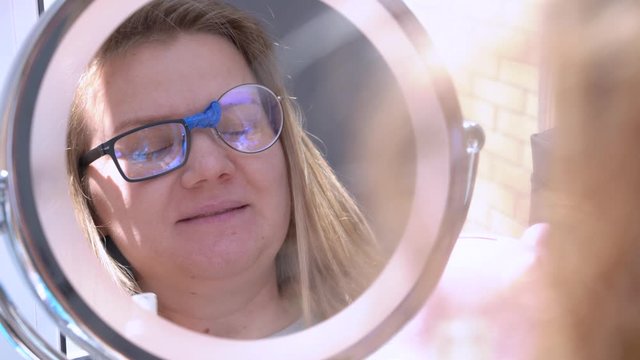 Crazy blonde girl in broken glasses for vision correction glued with duct tape from different lenses, looks at herself in the mirror and screams in surprise or fear from her appearance