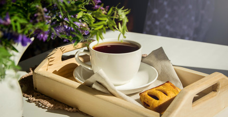 Summer breakfast. White cup with tea, butter biscuits on a wooden tray on a white table.