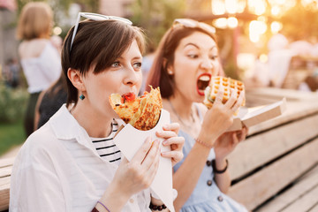 Two happy women friends relaxing in city park and eating modern street food cuisine. Healthy diet...