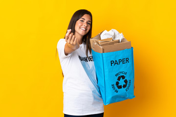 Young brazilian girl holding a recycling bag full of paper to recycle isolated on yellow background making money gesture