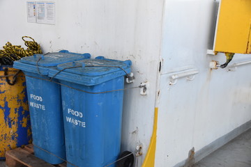 Blue plastic garbage bins for food waste firmly tight to the railings are situated behind superstructure of the cargo container vessel sailing through Atlantic ocean.