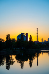 Obraz premium Production facility silo at sunset. Industrial complex located by the lake. Grain or other loose product storehouse silhouette on the horizon. Water reflection. 