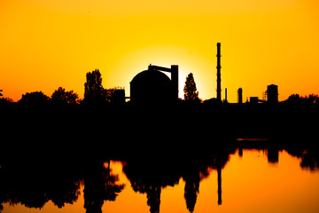 Production facility silo at sunset.  Industrial complex located by the lake. Grain or other loose product storehouse silhouette on the horizon. Water reflection. 