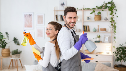 Big cleaning at house. Smiling husband and wife in aprons and rubber gloves with sprays in living room