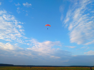 A skydiver with a bright orange parachute flies against a blue sky with white sparse clouds