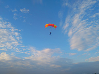 A skydiver with a bright orange parachute flies against a blue sky with white sparse clouds
