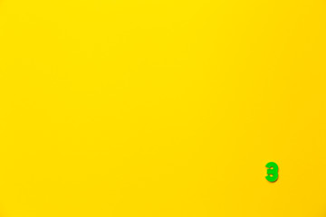 Number 3, three, the third of the plastic baby green toy number is located in the lower right corner on a yellow paper background. Banner on the topic of page numbering, timing or day in the calendar