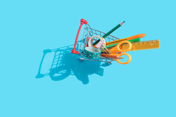 Back to school. Miniature supermarket trolley with stationery inside: scissors, pens, pencils, paper clips, ruler, tape. Blue background, top view,  flat lay.