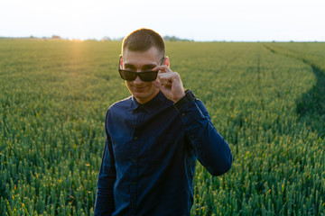 Close-up portrait of a handsome, attractive, smart, stylish, guy, straightens his sunglasses on the background of the field in the evening