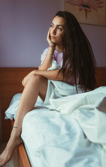 Pleased female in nightwear touching her face and looking at the window while sitting on soft bed in morning.